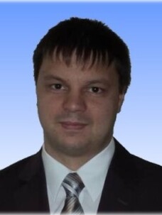 Astakhov mit kurzem dunklem Haar, im Anzug mit Krawatte, vor blauem Verlaufshintergrund.